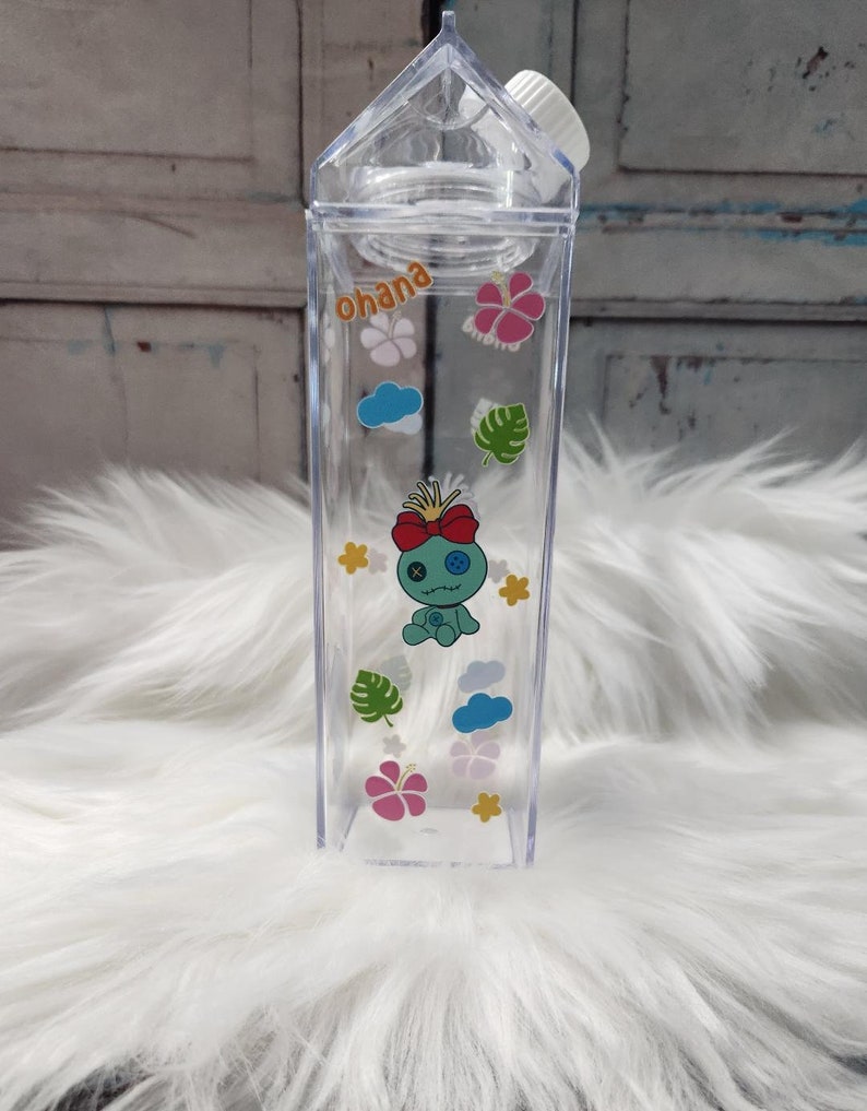 Puede incluir: Botella de agua de pl&aacute;stico transparente, con forma de cart&oacute;n de leche, con tap&oacute;n blanco. Decorada con pegatinas coloridas de un personaje de dibujos animados, flores, nubes y la palabra "ohana".