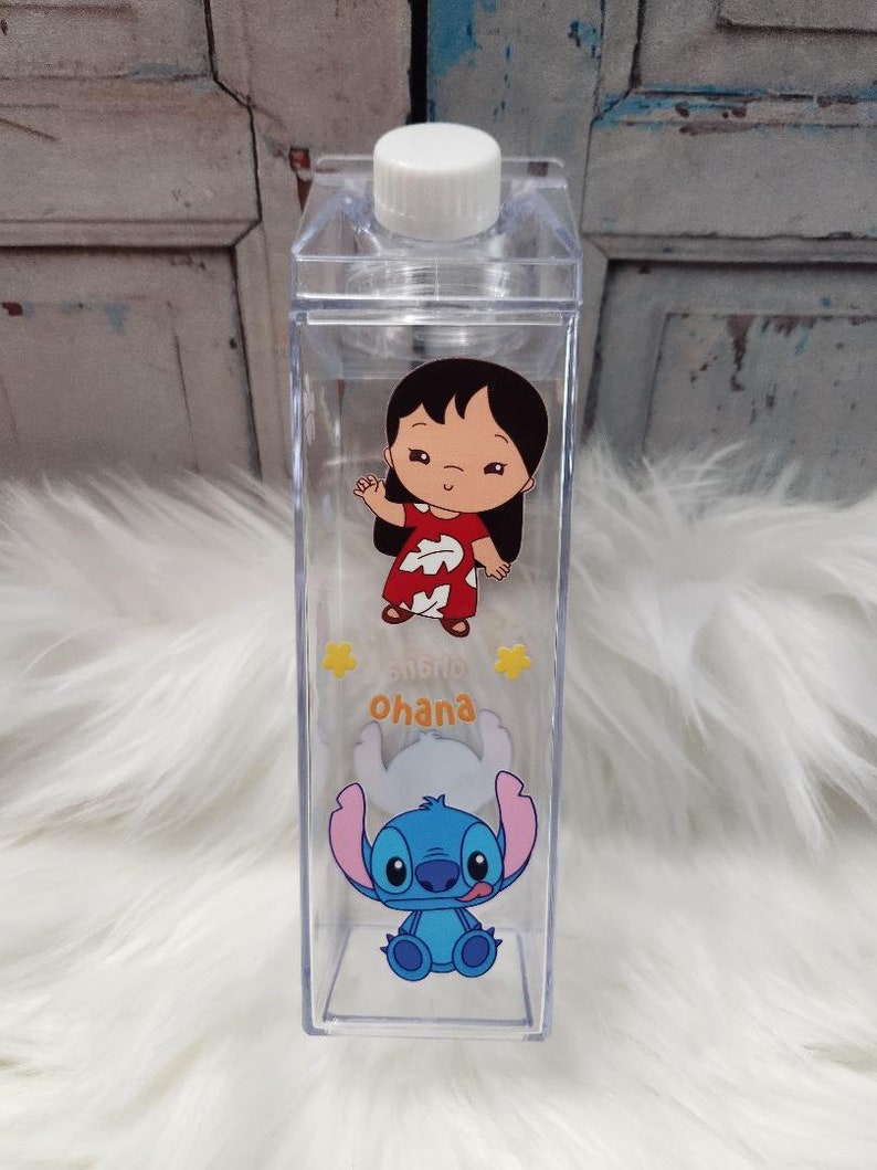 Puede incluir: Botella de agua de pl&aacute;stico transparente con tapa blanca. La botella presenta gr&aacute;ficos de dibujos animados de Lilo y Stitch, con la palabra "Ohana" en amarillo. La botella es rectangular y parece estar vac&iacute;a.