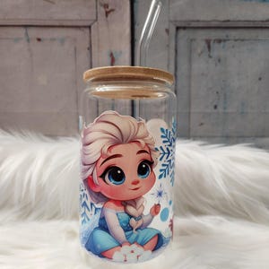 Peut inclure: Un bocal en verre transparent avec un couvercle en bambou et une paille. Le bocal est décoré d'une image colorée d'un personnage de dessin animé, Elsa de La Reine des Neiges, portant une robe bleue et avec des cheveux blonds. L'image est entourée de flocons de neige.