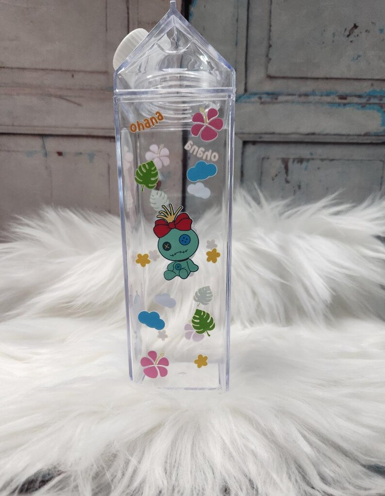 Puede incluir: Botella de agua de pl&aacute;stico transparente con forma de cart&oacute;n de leche, con tapa blanca. Decorada con gr&aacute;ficos de dibujos animados de una criatura azul, flores, nubes y la palabra "ohana".