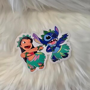 Puede incluir: Dos personajes de dibujos animados, Stitch y Lilo, est&aacute;n vestidos con faldas de hula y collares de flores. Stitch es azul con tres dedos en cada mano y lleva un collar de flores verde. Lilo lleva una camiseta roja y un collar de flores verde.