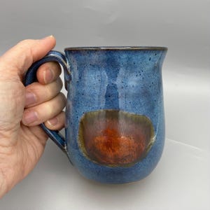 May include: A handcrafted ceramic mug with a blue glaze and a distinctive, earthy-toned design. The mug has a rounded form with a handle and a contrasting brown and orange accent. The mug is held by a hand.