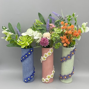 May include: Three cylindrical vases in blue, pink, and green, each decorated with a floral pattern. The vases contain an assortment of colourful artificial flowers and greenery. The vases are a decorative home accent, perfect for displaying flowers.