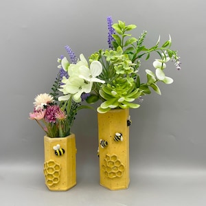 May include: Two yellow ceramic vases with a honeycomb design and bee accents. The vases hold colourful faux flowers and greenery. The vases are hexagonal and have a textured surface. The background is a solid grey.