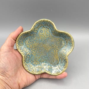 May include: A handmade, ceramic, flower-shaped dish with a detailed, embossed floral pattern. The dish has a metallic gold base with a turquoise patina, creating a unique, aged appearance. The dish is held in a hand.
