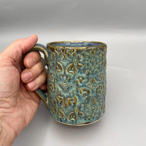 May include: A ceramic mug featuring a textured, raised damask-style pattern in shades of teal, blue, and metallic gold. The mug has a cylindrical body, a curved handle, and a smooth interior rim, showcasing a multi-toned glaze finish throughout.