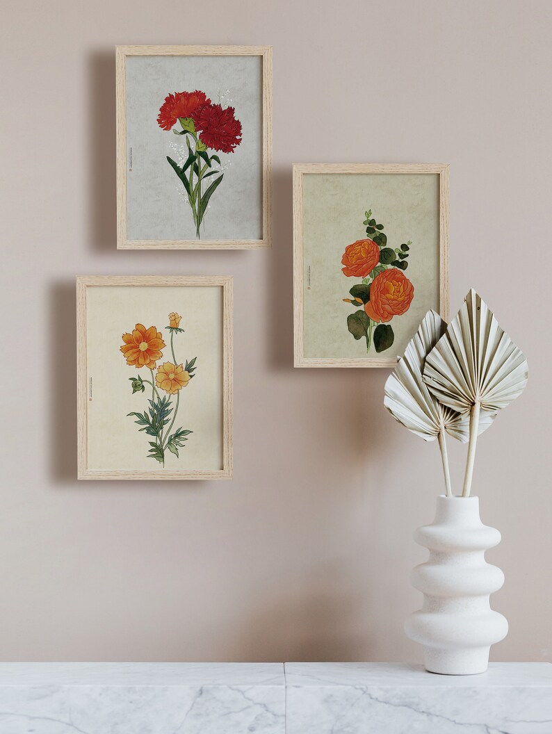 May include: Three framed botanical prints featuring different types of flowers. The prints are hung on a light beige wall. A white ceramic vase with a wavy design holds dried palm leaves.