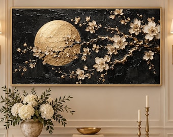 May include: A rectangular wall art piece featuring a textured black background with a large, circular gold moon and white floral branches extending across the canvas. The artwork is framed in a thin gold border and mounted on a wall.