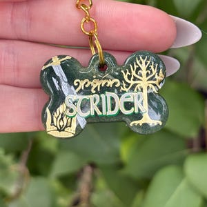 May include: A dog tag-shaped keyring with a gold-coloured ring and chain. The tag is dark green with gold accents and the name "SCRIDER" in white and green lettering. The tag features a tree and other designs.