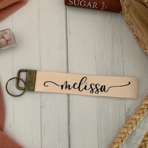 May include: A light pink canvas keychain with a black cursive font that reads "melissa". The keychain has a gold-colored metal ring and clasp.