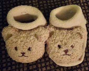 May include: A pair of baby booties in a light beige color. Each bootie features a teddy bear face design on the toe, with embroidered eyes, nose, and mouth. The top of the booties are a lighter shade of beige.