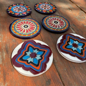 Op de afbeelding: Een verzameling keramische onderzetters met kleurrijke bloemmotieven. De onderzetters hebben verschillende patronen in blauw, rood, oranje en geel. De onderzetters zijn rond en hebben een donkerblauwe rand.