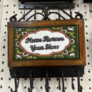 May include: A decorative wall-mounted sign with the text "Please Remove Your Shoes" on a green and orange tile. The sign is framed in wood and has several black hooks below for hanging items.