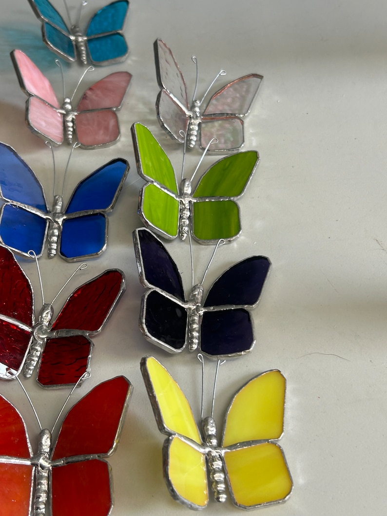 Pu&ograve; includere: Una collezione di farfalle in vetro colorato in vari colori, tra cui blu, verde, rosso, giallo e rosa. Le farfalle sono disposte in fila, con le ali spiegate.