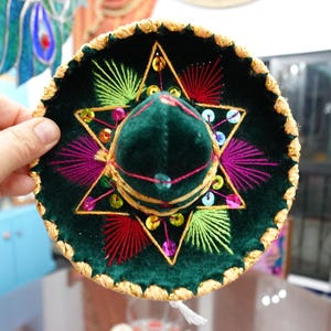 May include: A small, dark green sombrero with colorful embroidery and sequins. The hat features a star design with pink, yellow, and green accents, and a gold-colored trim. The hat is held by a hand.