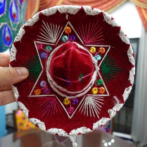 May include: A small, red sombrero with white embroidered trim and a central star design. The hat features colorful sequin accents and a velvet crown. This festive hat is a traditional Mexican accessory.