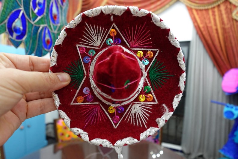 May include: A small, red sombrero with white embroidered trim and a central star design. The hat features colorful sequin accents and a velvet crown. This festive hat is a traditional Mexican accessory.