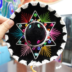 May include: A black sombrero with a white trim and colorful embroidery. The hat features a six-pointed star design with red, green, yellow, and pink thread. The center of the hat is black with a small black pom-pom.