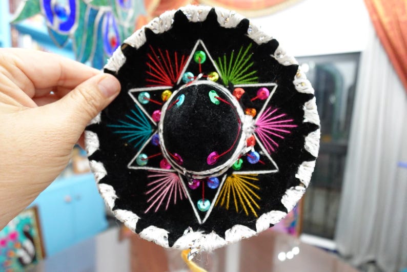 May include: A black sombrero with a white trim and colorful embroidery. The hat features a six-pointed star design with red, green, yellow, and pink thread. The center of the hat is black with a small black pom-pom.