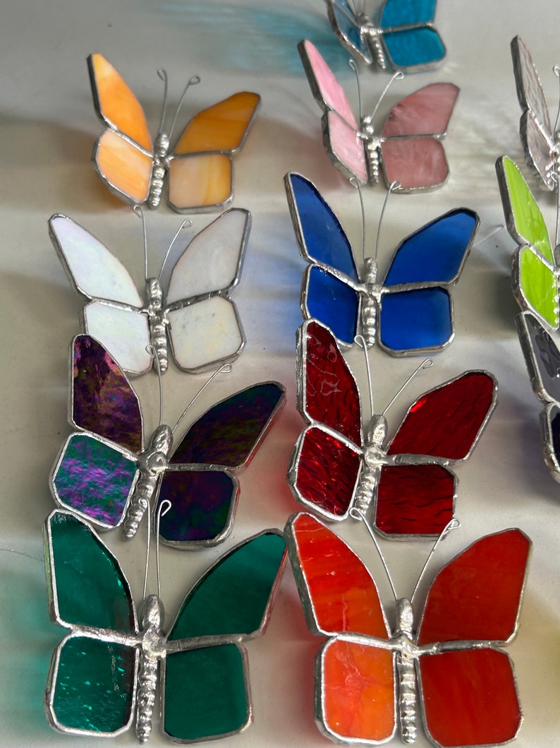 Pu&ograve; includere: Una collezione di farfalle in vetro colorato, tra cui arancione, bianco, blu, rosso, verde e viola. Le farfalle sono fatte di vetro e hanno una struttura in filo metallico.