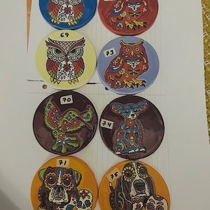 May include: Six round ceramic coasters with colorful designs. The coasters feature owls, hummingbirds, and dogs with sugar skulls. The coasters are numbered 68, 69, 70, 71, 72, 73, 74, and 75.