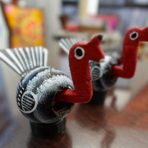 Puede incluir: Dos figuras de pájaros de madera pintadas a mano. Los pájaros tienen alas a rayas blancas y negras, cuerpos negros con detalles blancos y cabezas y picos rojos brillantes. Se asientan sobre pequeñas bases negras, mostrando un estilo de arte popular.