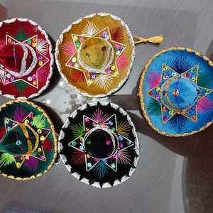 May include: A collection of miniature sombreros in various colors, including red, gold, blue, green, and black. Each hat is decorated with star-shaped embellishments and colorful embroidery, with a gold trim.