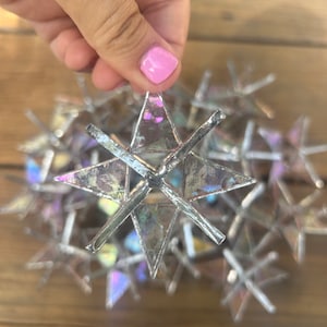 May include: A cluster of iridescent glass star ornaments, with one star held in a hand. The stars are made of multiple pieces of glass, creating a multifaceted, reflective surface.