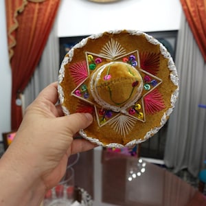 May include: A brown sombrero with colorful embroidery and a silver-colored trim. The hat features a star-shaped design with pink and white stitching, and small, colorful beads. A festive accessory.