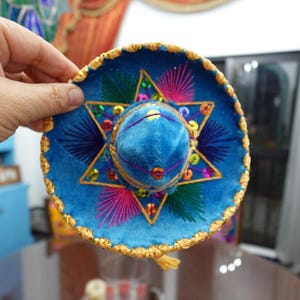 May include: A miniature blue sombrero with gold trim and colorful embroidery. The hat features a central star design with pink, green, and yellow accents. A small gold tassel hangs from the brim.