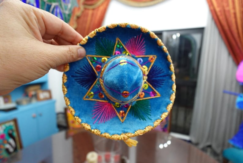 May include: A miniature blue sombrero with gold trim and colorful embroidery. The hat features a central star design with pink, green, and yellow accents. A small gold tassel hangs from the brim.