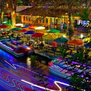 May include: A nighttime view of a river with brightly lit boats and a restaurant with colorful umbrellas and string lights. The restaurant has a patio with tables and chairs.
