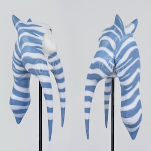May include: Two blue and white striped headpieces with long, pointed appendages. The headpieces are made of fabric and are mounted on black stands.