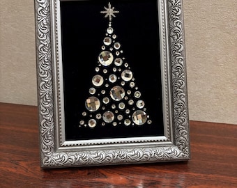 May include: A framed Christmas tree art piece. The artwork features a black background with a tree design made of various sized clear, sparkling crystals. The frame is silver with ornate detailing. A star tops the tree.