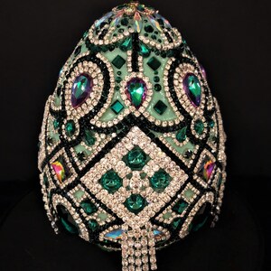 May include: A green egg-shaped object covered in rhinestones and other jewels. The egg is decorated with a geometric pattern and has a fringe of rhinestones hanging from the bottom.