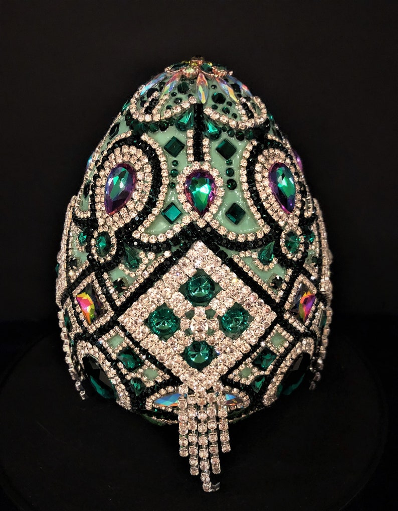 May include: A green egg-shaped object covered in rhinestones and other jewels. The egg is decorated with a geometric pattern and has a fringe of rhinestones hanging from the bottom.