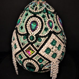 May include: A green egg-shaped object covered in rhinestones and green and black jewels. The egg is sitting on a black surface.