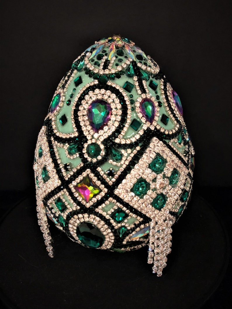 May include: A green egg-shaped object covered in rhinestones and green and black jewels. The egg is sitting on a black surface.