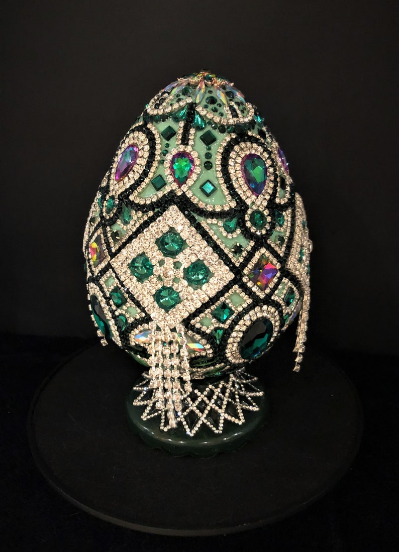 May include: A green egg-shaped decorative object covered in rhinestones and beads. The egg is on a stand and has a geometric design.
