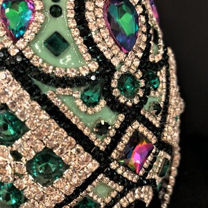 May include: A green egg-shaped object covered in rhinestones and small green and iridescent gems. The object is decorated with a black and white geometric pattern.