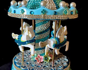 May include: A decorative miniature carousel in shades of turquoise and silver. It features white horses with gold detailing, wearing blue jeweled collars. The roof and base are adorned with rhinestones and pearls, creating a sparkling effect.