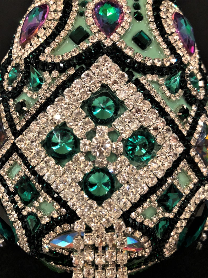 May include: A green and black egg-shaped ornament covered in rhinestones and emerald green jewels. The ornament has a geometric pattern.