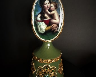 May include: A green and gold musical egg with a painted image of the Madonna and Child. The egg is decorated with gold trim and green jewels.