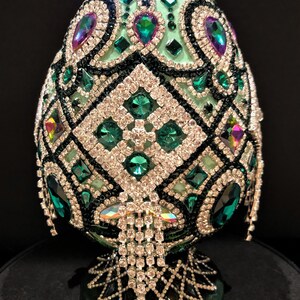 May include: A green egg-shaped decorative object covered in rhinestones and emerald green jewels. The egg is on a green stand with a diamond pattern.