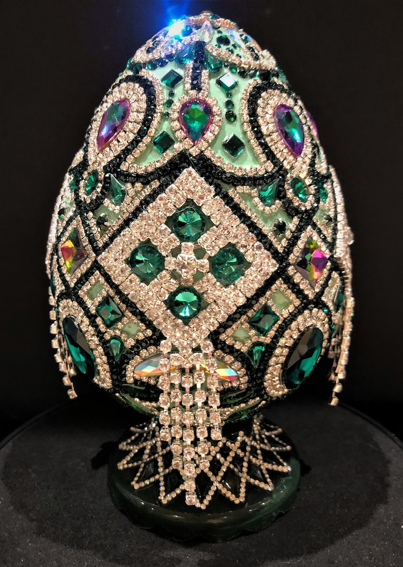 May include: A green egg-shaped decorative object covered in rhinestones and emerald green jewels. The egg is on a green stand with a diamond pattern.