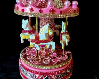 May include: A pink and gold carousel music box. The carousel features white horses with gold detailing, a pink and gold roof, and is embellished with rhinestones and pearls. The base is also pink and decorated with rhinestones. A decorative item.