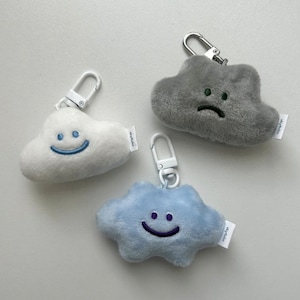 May include: Three plush cloud keyrings with different facial expressions. One cloud is grey with a frowning face, one is white with a smiling face, and one is light blue with a smiling face. Each keyring has a silver metal ring and a white tag with the text "skyfelia".