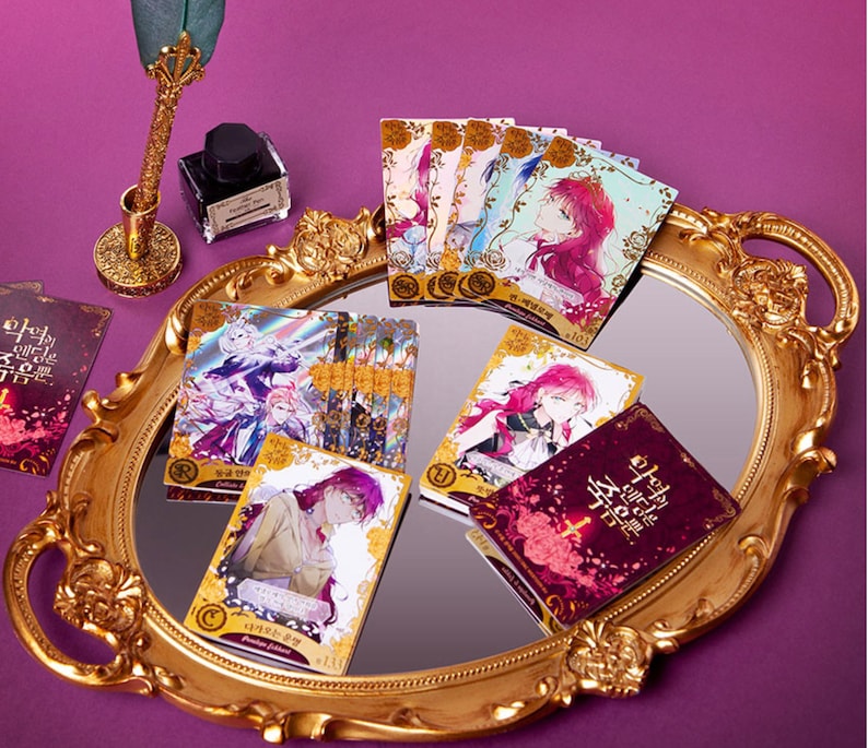 May include: A collection of trading cards featuring illustrations of characters from the Korean webtoon "The Remarried Empress". The cards are displayed on a gold-colored tray with a mirror surface. The cards have a holographic finish and feature the text "The Remarried Empress" in Korean.