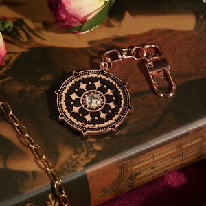 May include: A rose gold-colored keychain charm with a circular, ornate design. The charm features a black center with a clear crystal and intricate detailing. A gold chain and a rose are also visible.