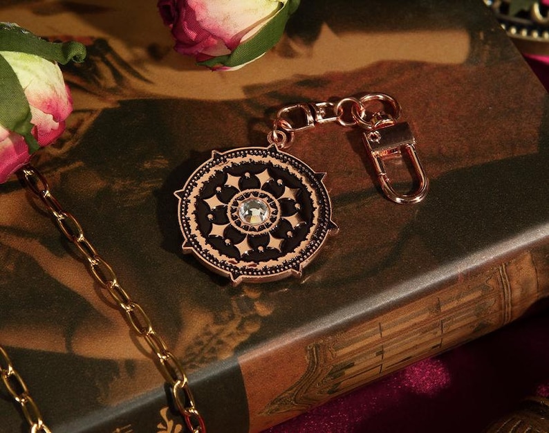 May include: A rose gold-colored keychain charm with a circular, ornate design. The charm features a black center with a clear crystal and intricate detailing. A gold chain and a rose are also visible.